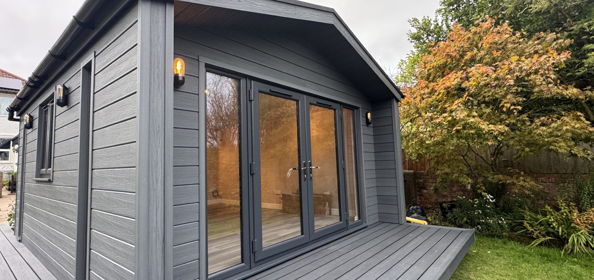 Modern grey wooden cabin with large glass doors and a deck, surrounded by greenery.